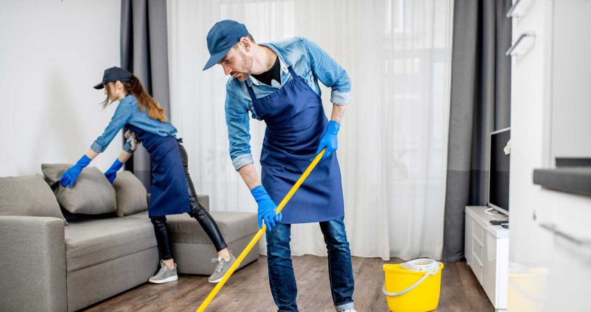 Two professional cleaners in blue uniforms and gloves tidying up a modern living room; a man mopping the floor with a yellow mop and a woman arranging cushions on a gray sofa. A yellow cleaning bucket is visible on the floor.